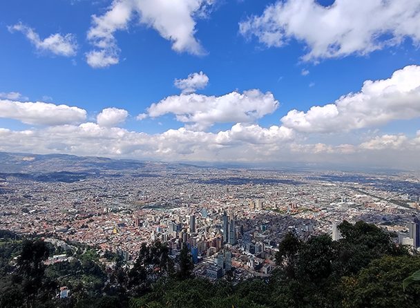 Aerial view of a sprawling cityscape under a bright blue sky with clouds. The city is surrounded by distant mountains and greenery in the foreground, perfect for those considering Bogotá city tours.