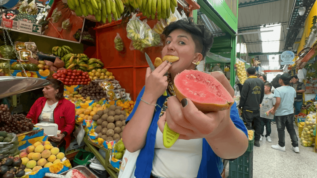 A person in a bustling Bogota market delights in eating sliced guava while holding another half and playfully showing it to the camera. Surrounded by an array of vibrant fruits, this scene captures the lively essence of one of Bogota's most charming tourist attractions.