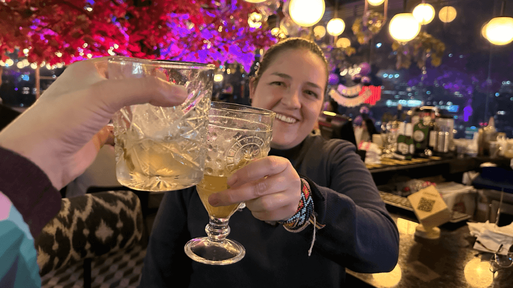 Two people clink glasses in a festive, warmly lit setting, surrounded by colorful decorations reminiscent of Bogota tourist attractions.