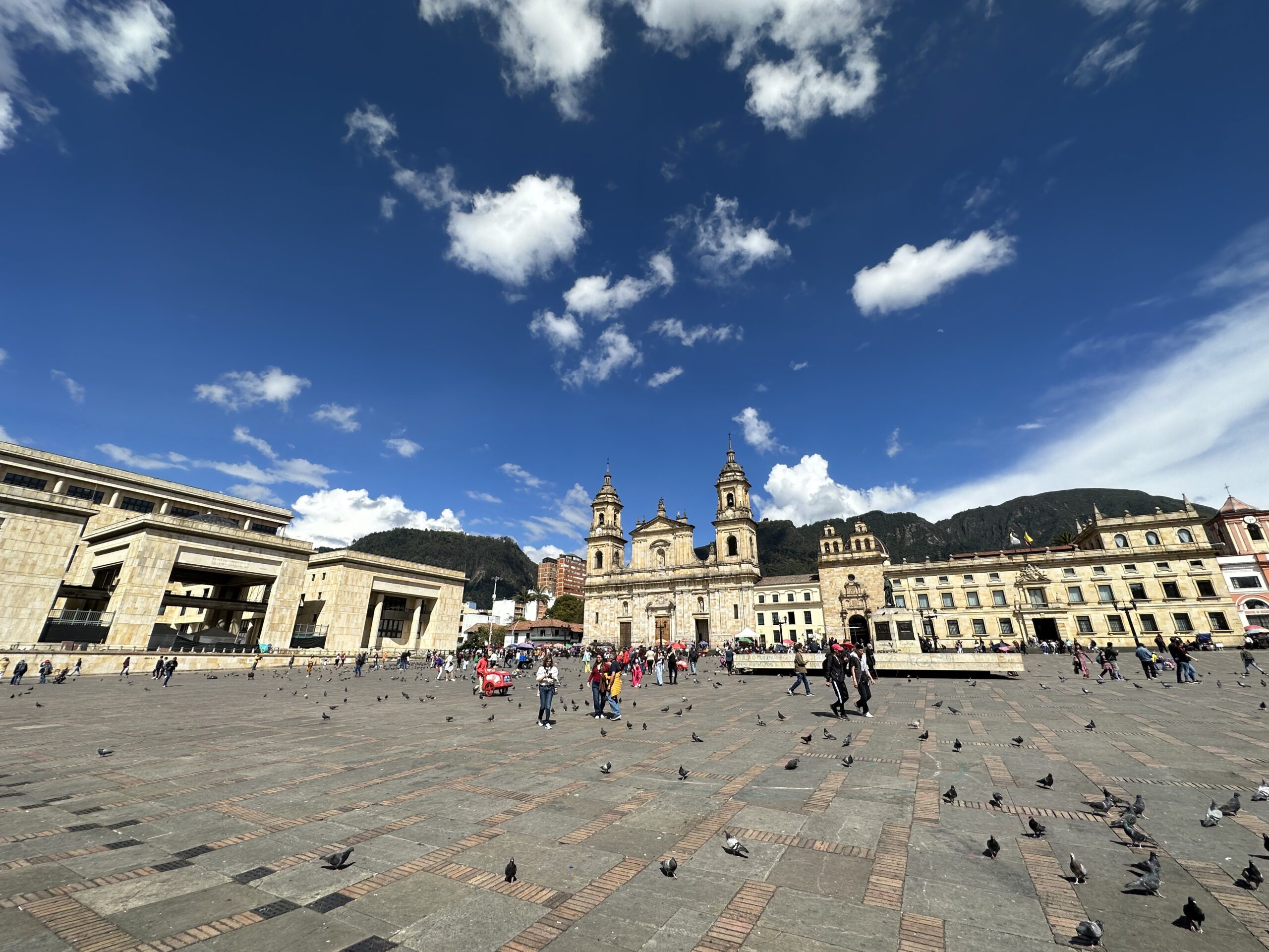 A wide view of a large cobblestone plaza with scattered people and pigeons, surrounded by historic buildings and a mountainous backdrop under a partly cloudy sky, creates the perfect setting for Bogota city tours.
