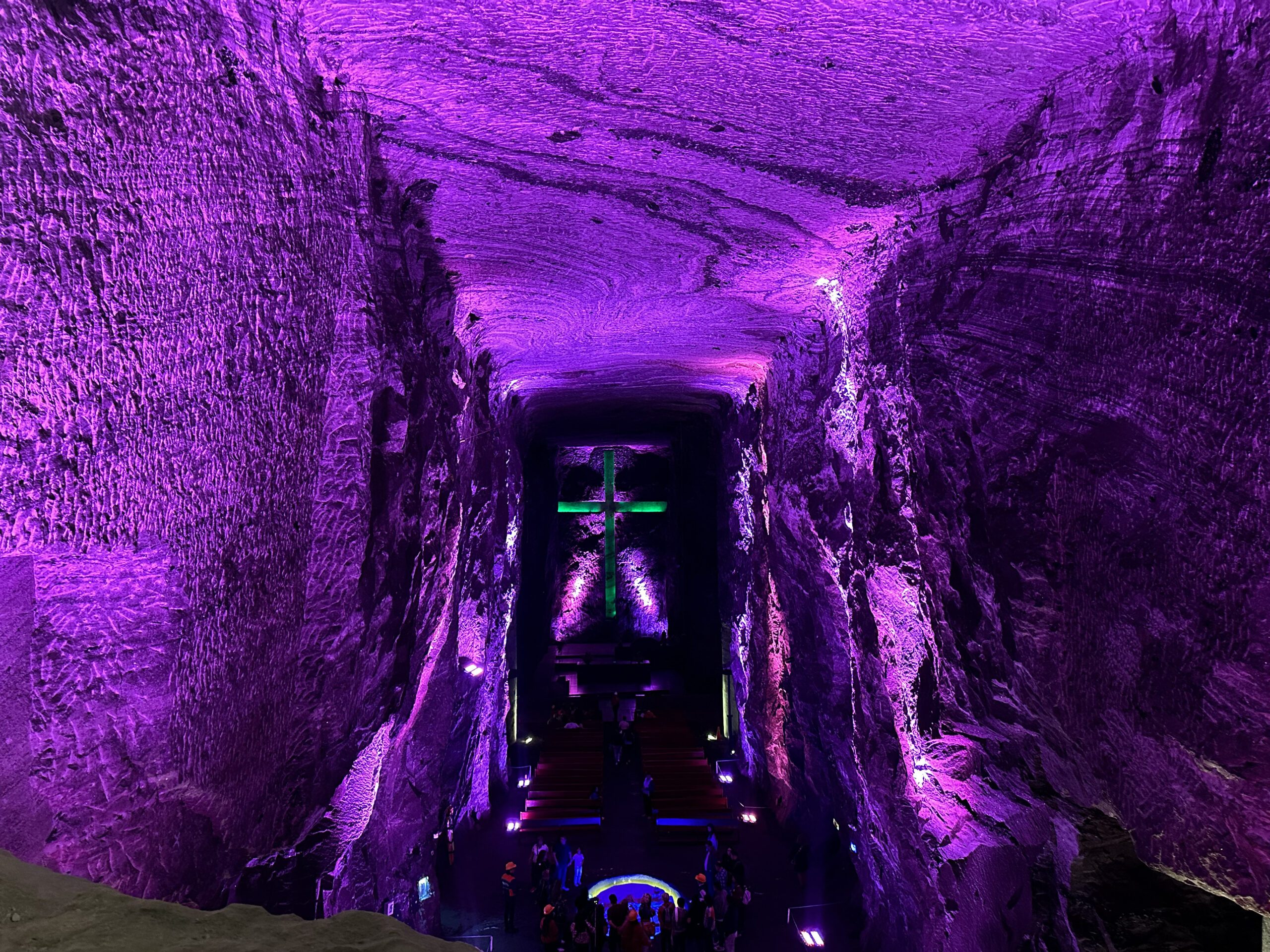 A large underground cavern lit with purple lights features a cross-shaped structure at the far end, with people gathered at the bottom—an unforgettable addition to your Bogotá city tours.
