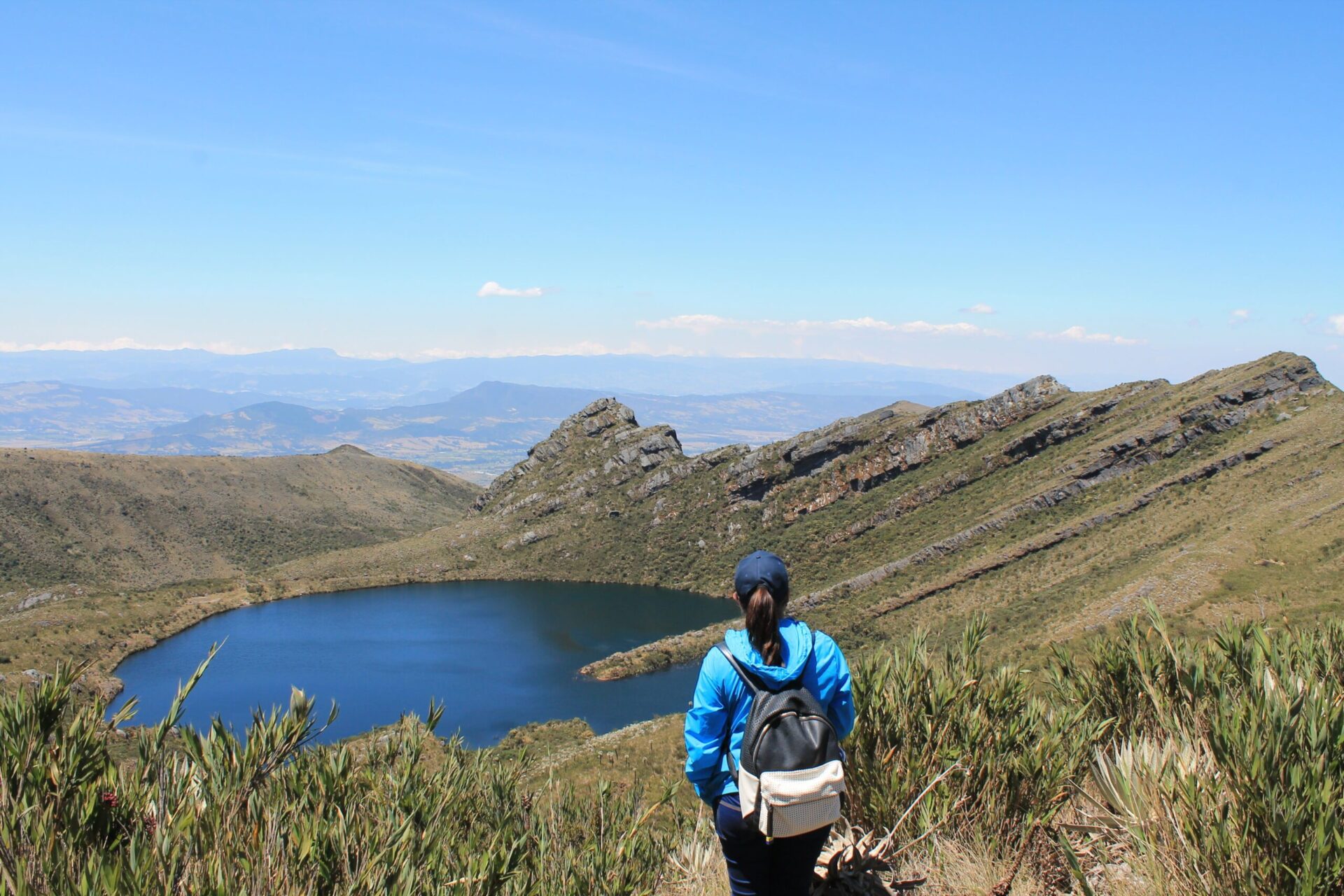 Chingaza: A person wearing a blue jacket and backpack is standing on a grassy hillside overlooking a lake surrounded by mountains under a clear blue sky, possibly reminiscing about their recent Bogotá city tours.