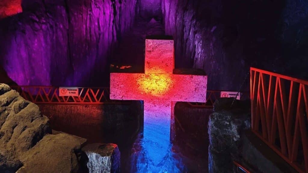 Explore an illuminated cross sculpture in a dimly lit cavern near Bogotá, where colored lights in purple, red, and blue create an enchanting atmosphere. Perfect for day trips, this cultural wonder is surrounded by rugged rocks and safety signs for your adventure.