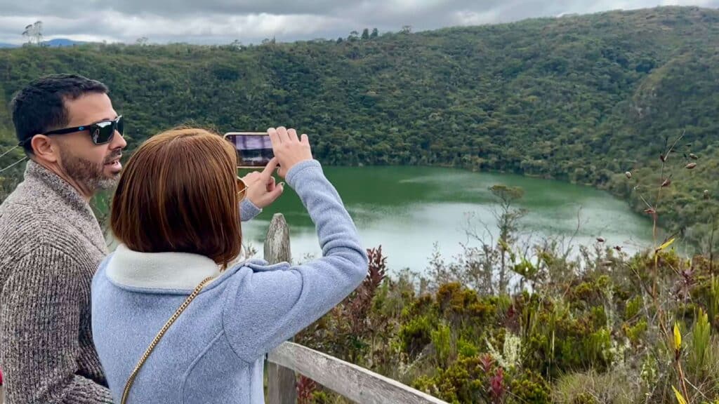 Two people stand at a wooden railing, capturing the essence of a green lake surrounded by trees, reminiscent of day trips around Bogota. One person is taking a photo with a phone as the overcast sky casts a serene ambiance over this natural wonder.