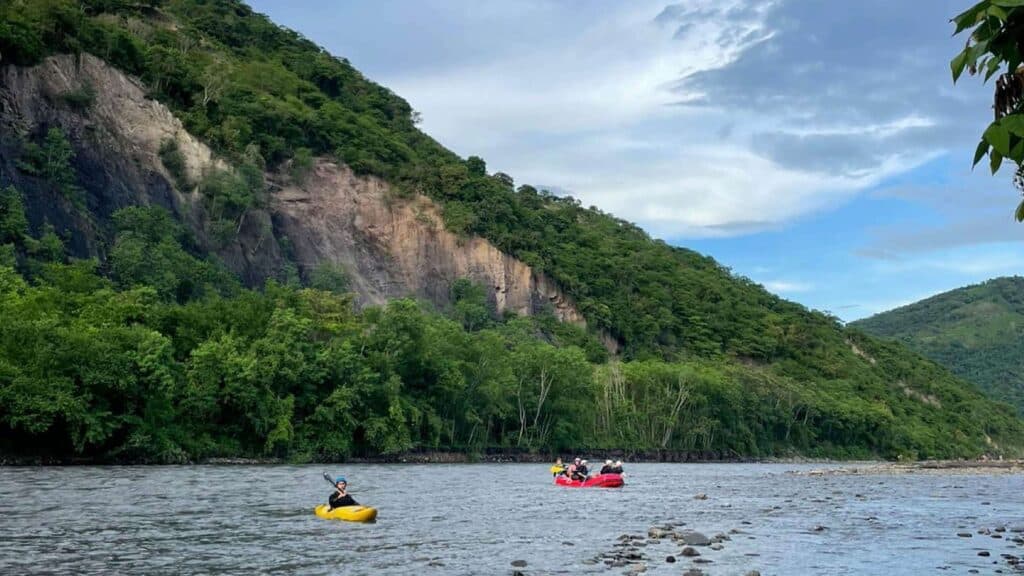 Kayakers embark on scenic day trips down a river near Bogotá, navigating waters framed by lush green hills and a rocky cliff under a partly cloudy sky, discovering the region's natural beauty alongside its cultural wonders.