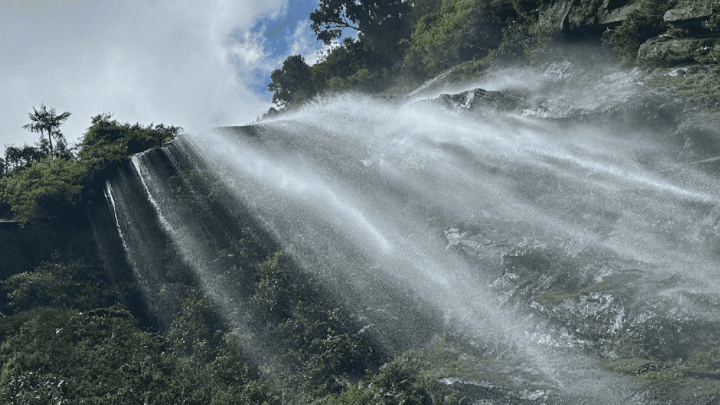 A waterfall cascades down a rocky cliffside, with water spray illuminated by sunlight. Sparse greenery surrounds the rocks under a partly cloudy sky, offering a breathtaking scene perfect for Day Tours from Bogota.