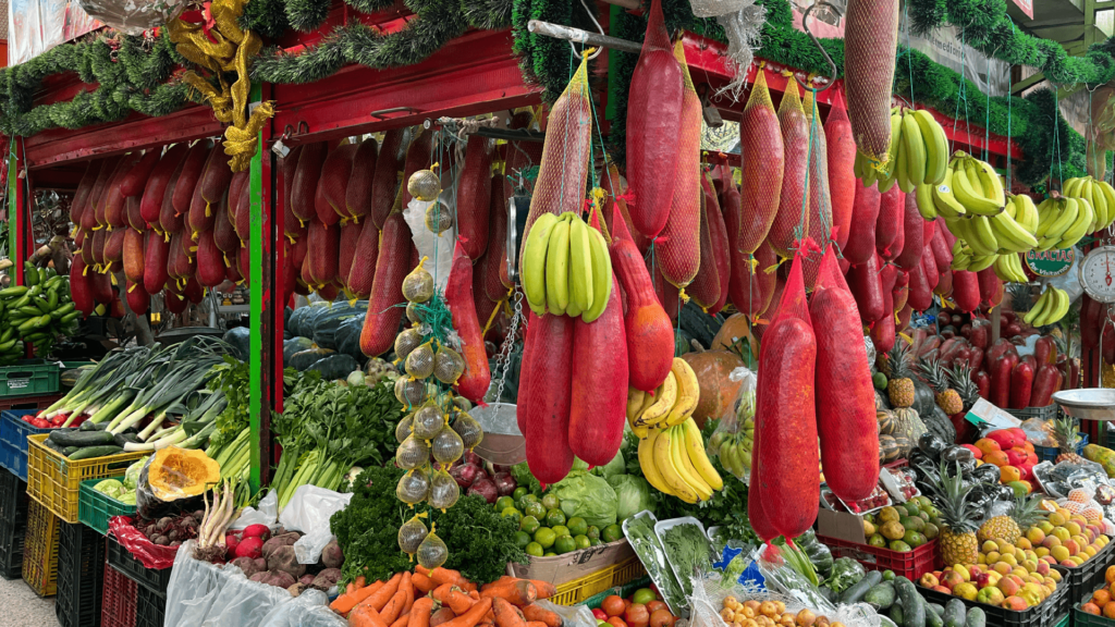 A market stall in Bogota showcases a variety of hanging cured meats and bananas, surrounded by fresh produce like carrots, lettuce, tomatoes, and squash—an essential stop on any day tour exploring the vibrant local flavors.