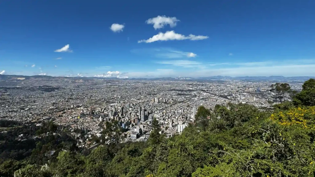 Aerial view of a sprawling cityscape under a clear blue sky, surrounded by green hills and scattered clouds—a perfect snapshot for those considering day tours of Bogota.