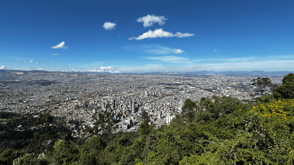 Aerial view of a sprawling cityscape under a clear blue sky, surrounded by green hills and scattered clouds—a perfect snapshot for those considering day tours of Bogota.