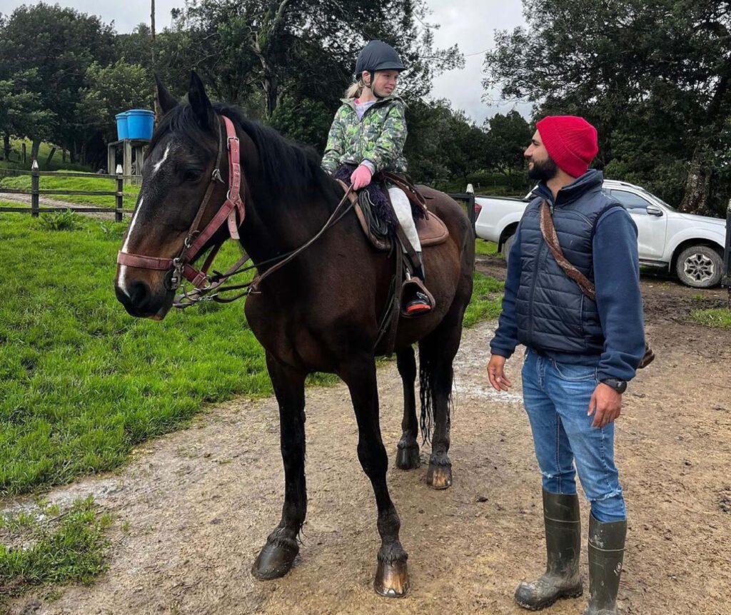 A child sits atop a brown horse, donning a helmet and jacket. Beside them, an adult in a red beanie and rain boots smiles warmly. Embrace the spirit of adventure with Family Friendly Tours Bogota, set against a backdrop of trees and a white truck.