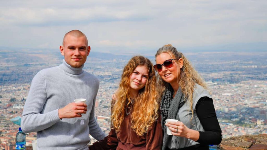 Three people stand together outside, holding cups. A man in a sweater and a woman in sunglasses flank a young girl with long hair. As they enjoy their drinks, the vibrant cityscape of Bogota serves as a perfect backdrop for their family-friendly exploration.