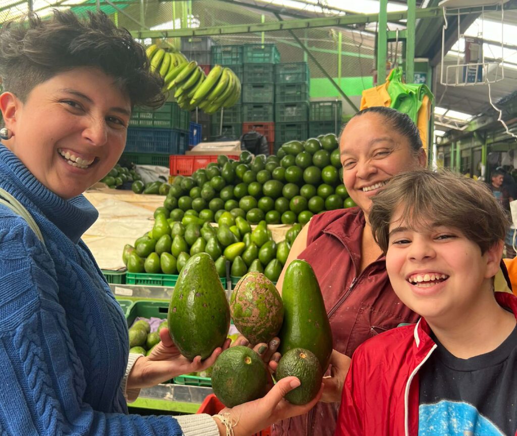 Three people smiling while holding avocados at a market stall, embodying the spirit of Family Friendly Tours Bogota. In the background, more avocados and bananas hang above, creating a vibrant atmosphere.