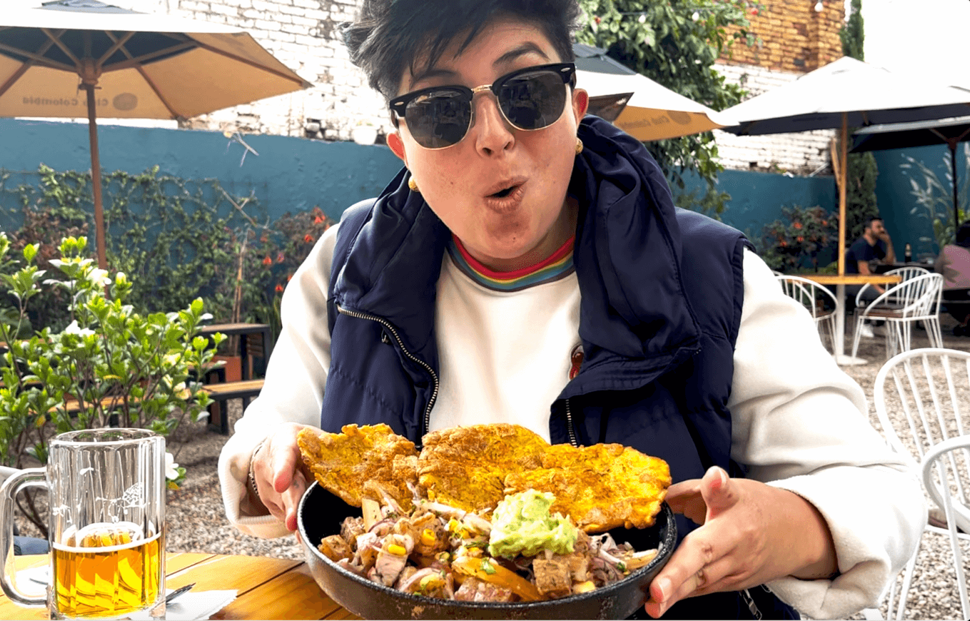Food Tour in Bogotá: Person wearing sunglasses and a vest, sitting outdoors in Bogotá, holding a plate of mixed grilled food with plantains and guacamole, with a drink on the table. Perfect for those looking for things to do in Bogotá after enjoying one of the many city tours.