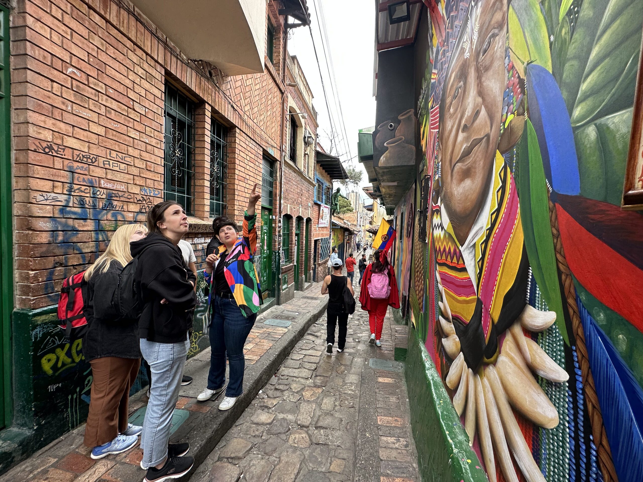 Tourists stand on a narrow cobblestone street, admiring a large, colorful mural on a brick wall. One person points out details of the mural while others observe. The street has graffiti and buildings—one of many captivating sights on Bogotá city tours.