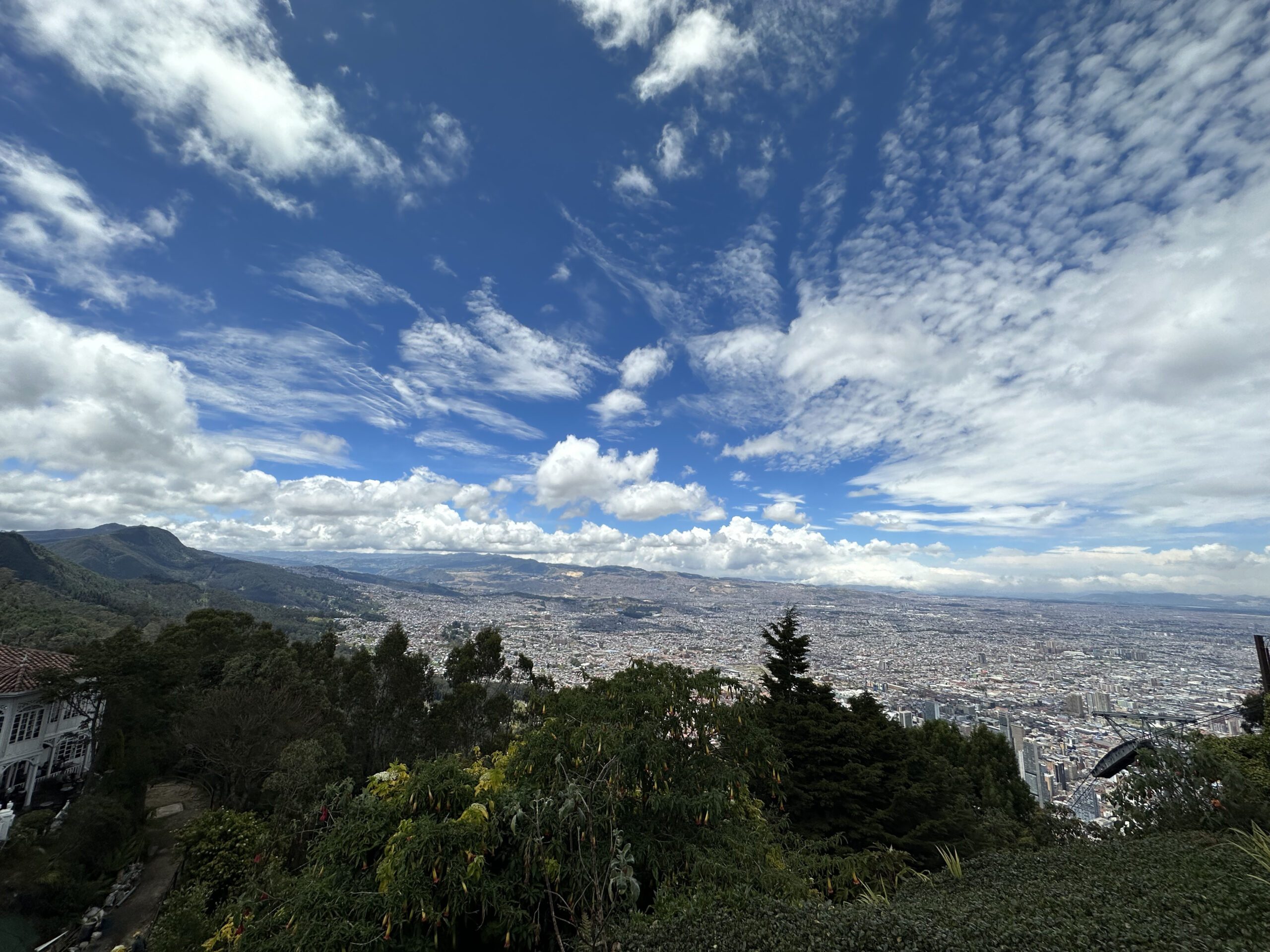 A panoramic view of a cityscape under a bright blue sky with scattered clouds, taken from a vantage point overlooking lush greenery, is one of the many highlights of Bogotá city tours.