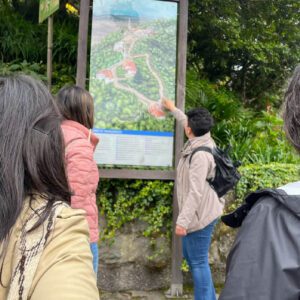 A group of people on a Bogotá day tour gathers around a large outdoor map. One person points at the map, surrounded by lush trees and vibrant plants in the background.