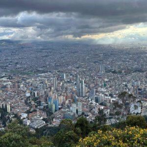 Aerial view of a sprawling cityscape with tall buildings and cloudy skies, surrounded by green hills—perfect for a Bogotá day tour.