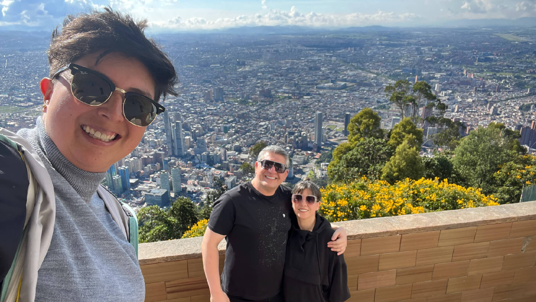 Three people capturing a selfie above the vibrant cityscape of Bogota, with lush greenery and bursts of yellow flowers in the foreground, perfectly capturing their day tour adventure.