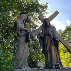 In a serene garden with lush greenery and a clear blue sky, a statue of Jesus carrying a cross is joined by another figure. This captivating scene is reminiscent of the spiritual stops often explored on a Bogota day tour.