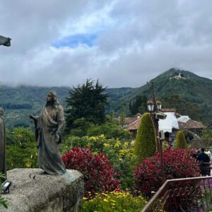 Amid a lush garden in the backdrop of majestic mountains under a cloudy sky, a statue of Jesus on the cross stands with two figures below. As part of a Bogota day tour, visitors stroll along the nearby pathway, soaking in the serene atmosphere.