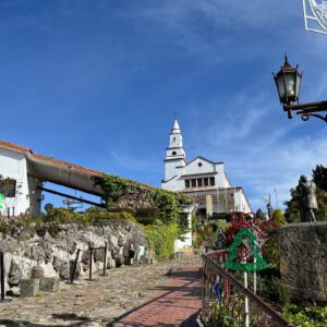 A pathway leads up to a white church with a tall tower, reminiscent of those seen on a Bogota day tour. The scene includes people walking, festive decorations, and clear blue skies.