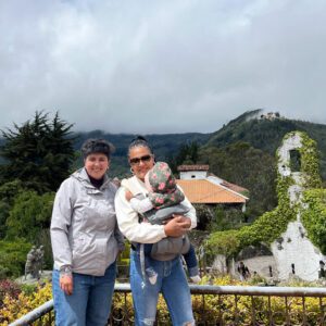 Two people and a baby enjoy a Bogota day tour, standing before a scenic view with lush trees, a cloudy sky, and a building partially draped in ivy.