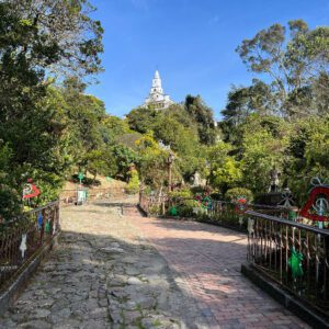 A stone pathway leads uphill through a lush garden towards a white building with a tower under the clear blue sky, offering views reminiscent of a Bogota day tour.