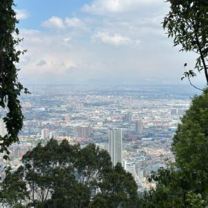 Experience a Bogota day tour with a cityscape view framed by trees, revealing numerous buildings under a partly cloudy sky.