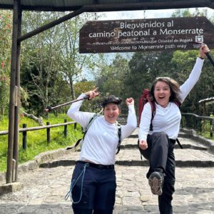 Two people joyfully pose under a "Welcome to Monserrate path" sign, capturing the essence of a Bogota day tour. Clad in hiking gear and gripping trekking poles, they embody the spirit of adventure.