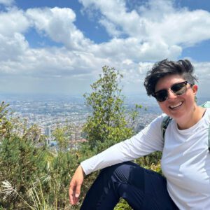 A person with short hair and sunglasses smiles while enjoying a Bogota day tour, sitting on a hill with the cityscape visible in the background under a partly cloudy sky.