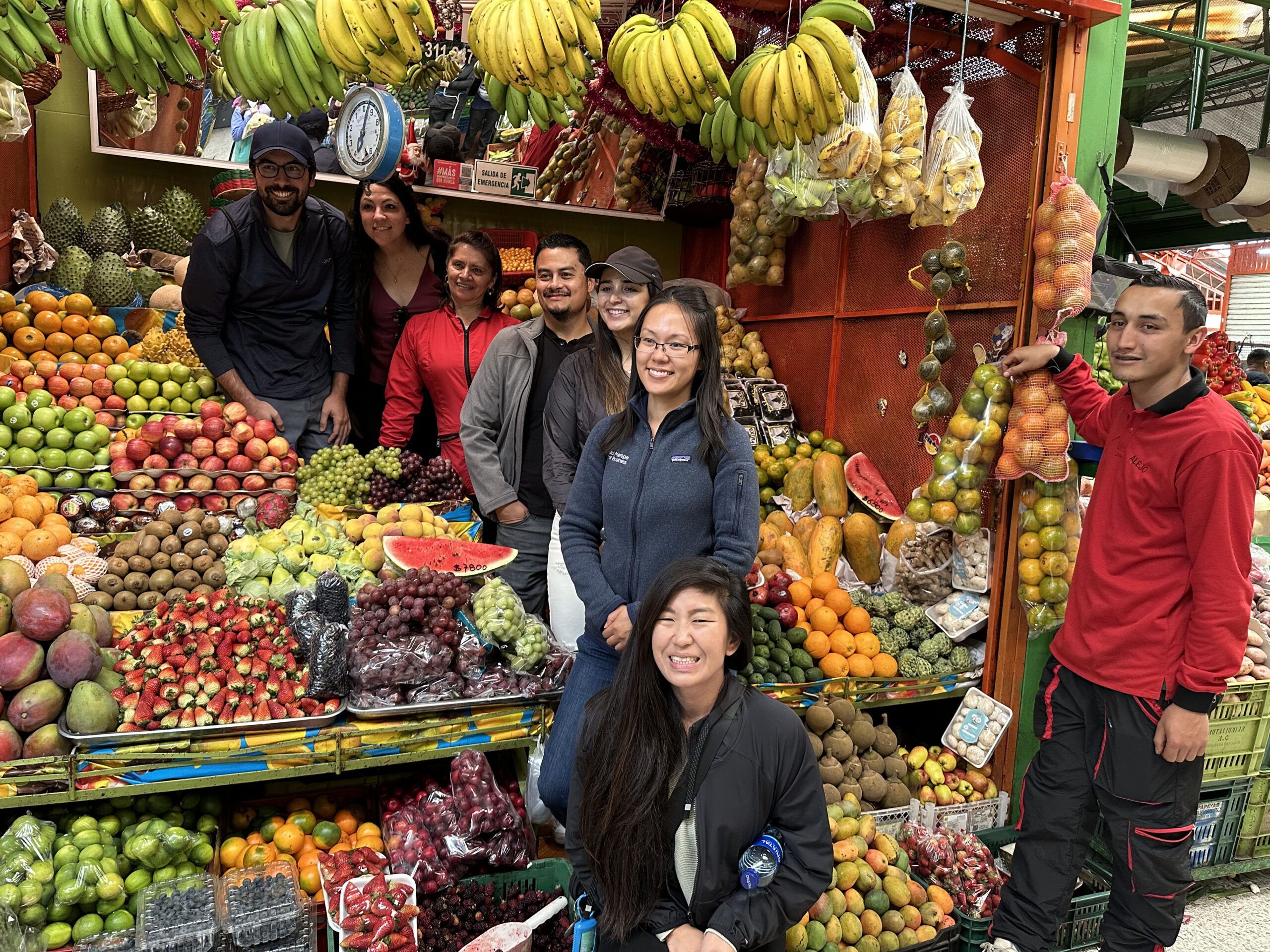 A group of people posing for a photo in front of a colorful fruit stand at a market in Bogotá, surrounded by various fruits such as bananas, grapes, apples, and oranges—a vibrant highlight of Bogotá city tours.