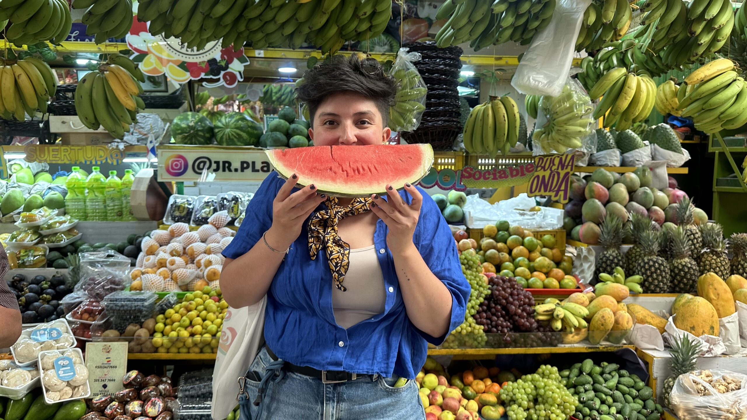 A person stands in front of a colorful fruit market display in Bogotá, holding and taking a bite of a large slice of watermelon, while wearing a blue shirt and jeans—a perfect snapshot from one of the vibrant tours in Bogotá.