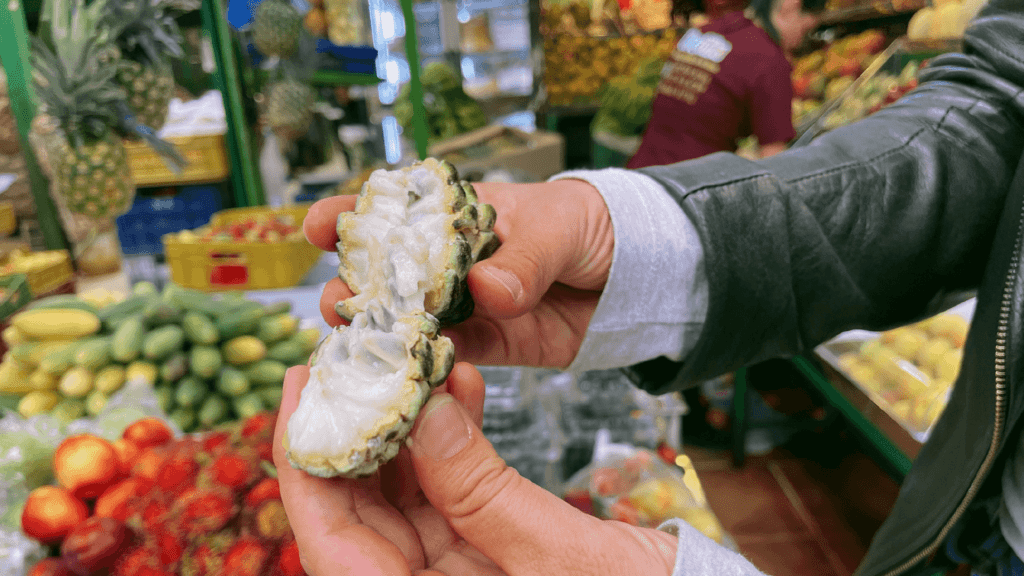 In the bustling Paloquemao Fruit Market of Bogota, a person stands amidst a vibrant array of fruits and vegetables, holding an opened tropical fruit with white flesh, adding a touch of magic to the bustling scene.