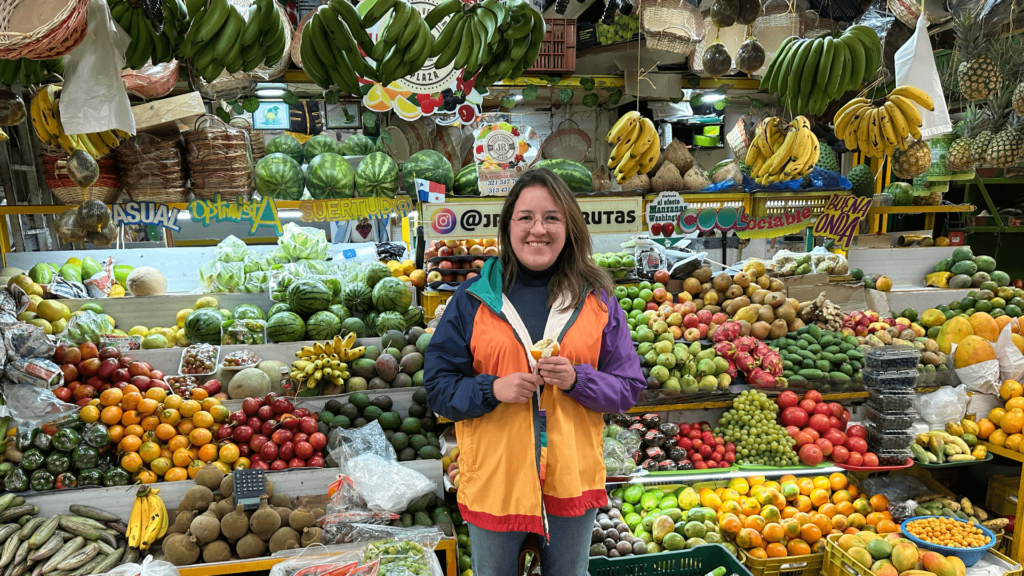 A person stands smiling in front of a colorful fruit stand at Bogota's Paloquemao Fruit Market, wearing a vibrant jacket. With an array of fruits like bananas, apples, and watermelons displayed around, the scene captures the market's sheer magic.