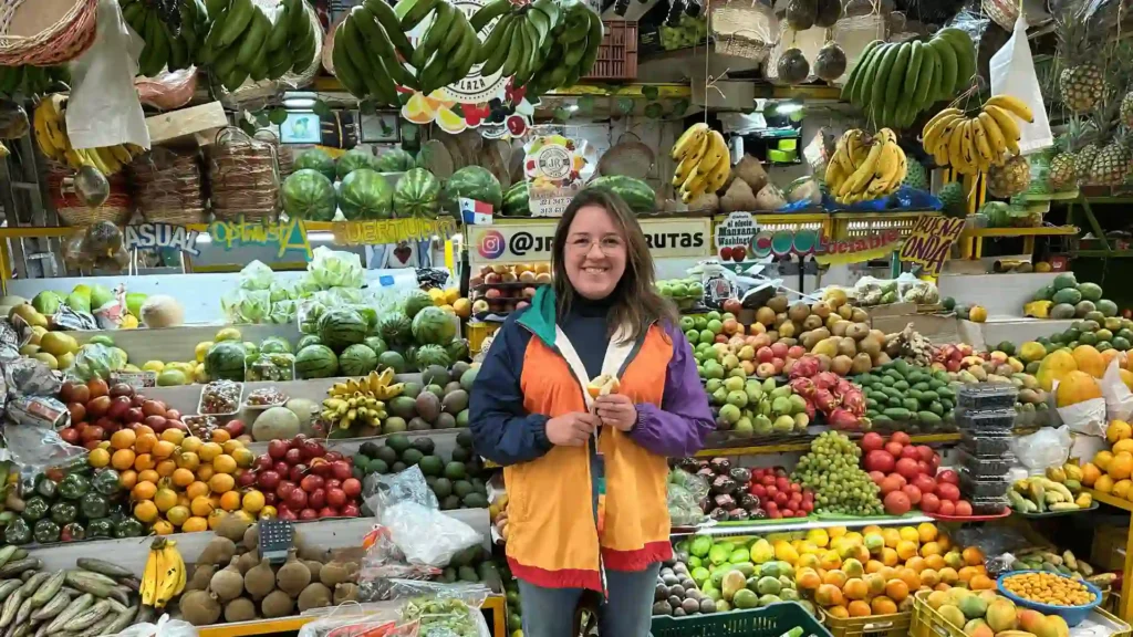 A person stands smiling in front of a colorful fruit stand at Bogota's Paloquemao Fruit Market, wearing a vibrant jacket. With an array of fruits like bananas, apples, and watermelons displayed around, the scene captures the market's sheer magic.