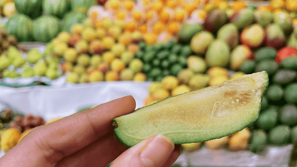 Close-up of a hand holding a bitten slice of green fruit with a market stall of various fruits in the blurred background at Paloquemao Fruit Market, Bogota.