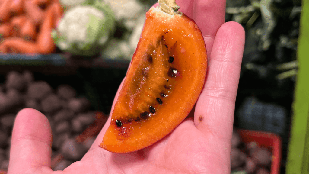 A hand holding a sliced piece of orange fruit with black seeds visible, seemingly infused with magic, captures the vibrant essence of Bogota’s Paloquemao Fruit Market.