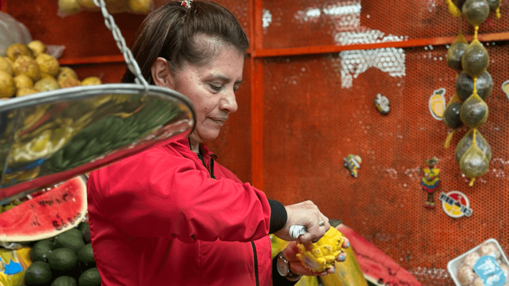 At the bustling Paloquemao Fruit Market in Bogotá, a woman in a red jacket performs a small act of magic as she skillfully peels a fruit, surrounded by an array of vibrant fruits and vegetables.