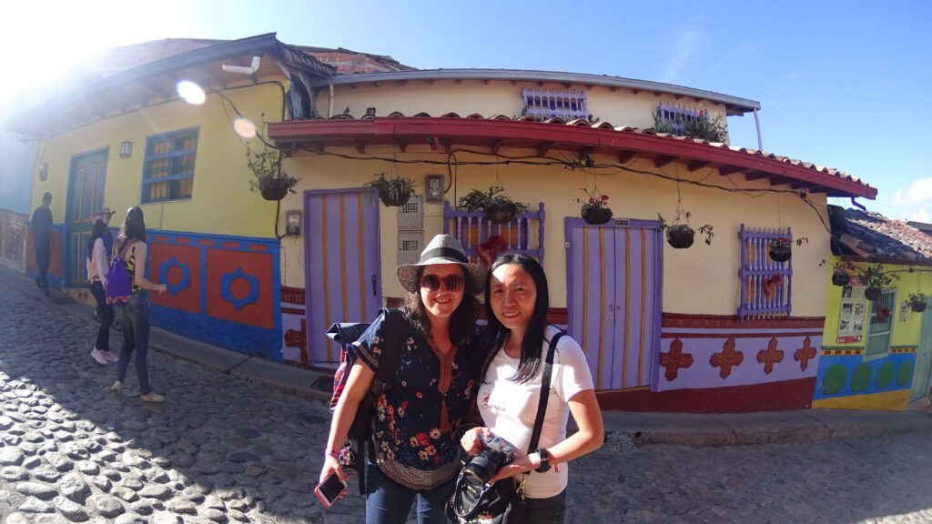 Two people stand on a cobblestone street in front of a colorful building with hanging plants and bright doors, capturing memories. One holds a phone, the other a camera, as they explore like locals might on solo tour packages.