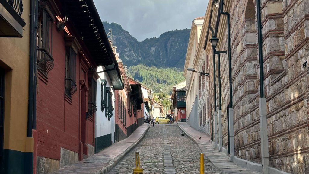 A one-day itinerary in Bogota reveals a charming cobblestone street lined with historic buildings, leading to a breathtaking mountain view under a cloudy sky.