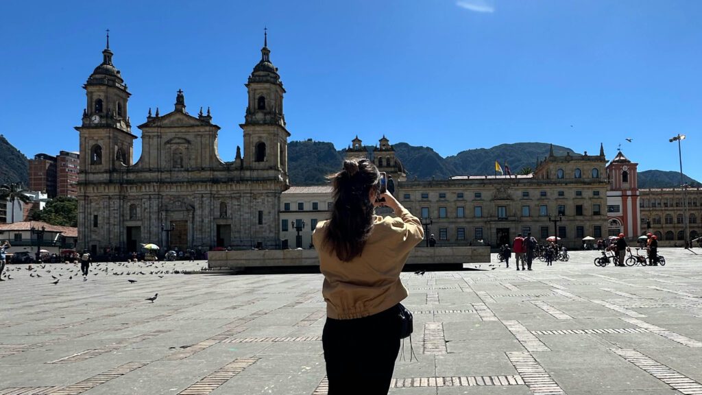 A person capturing a photo of a historic cathedral and surrounding buildings in a sunny plaza, with a mountainous backdrop, perfect for your one day itinerary of Bogotá's top things to see.