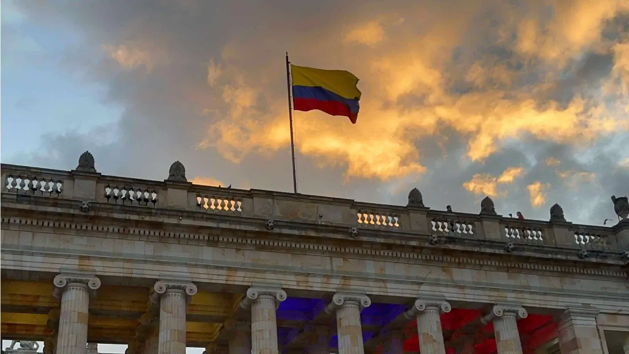 The Colombian flag waves atop a neoclassical building with columns at sunset, creating a perfect backdrop for those on the Candelaria Walking Tour. The dramatic sky enhances the scene, making it a must-visit spot for any traveler exploring Bogotá's rich history.