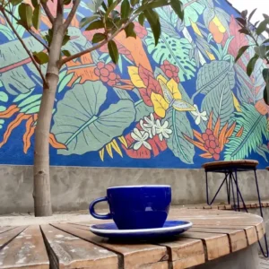 A blue coffee cup and saucer sit on a wooden table in front of a vibrant mural featuring large leaves and flowers. A small tree and a metal stool are also present, evoking the charm found in the hidden gems often highlighted on tours in Bogotá.