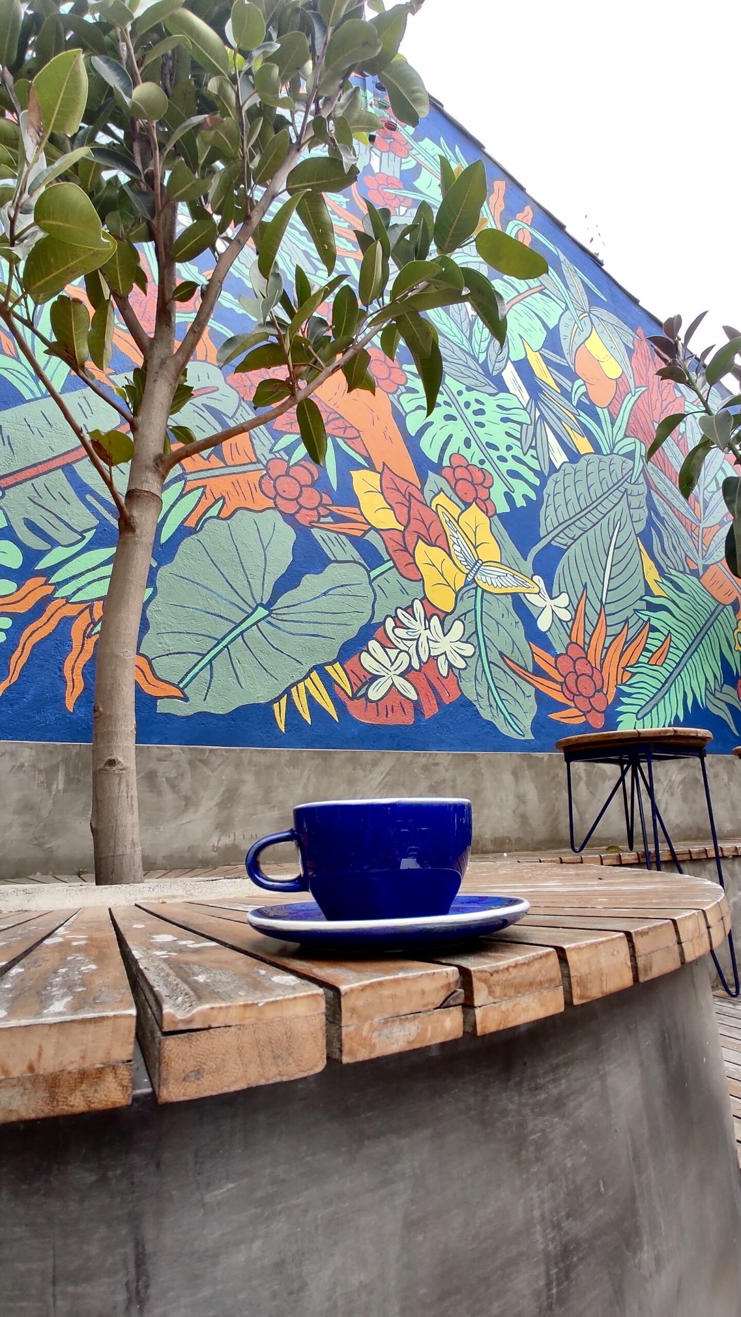 A blue coffee cup and saucer sit on a wooden table in front of a vibrant mural featuring large leaves and flowers. A small tree and a metal stool are also present, evoking the charm found in the hidden gems often highlighted on tours in Bogotá.