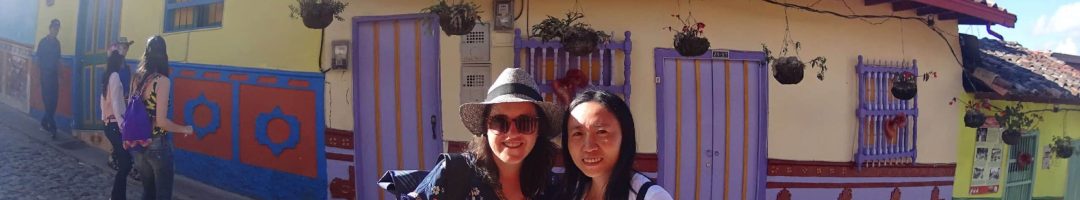 Two people stand on a cobblestone street in front of a colorful building with hanging plants and bright doors, capturing memories. One holds a phone, the other a camera, as they explore like locals might on solo tour packages.