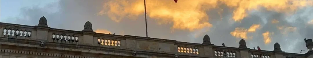 The Colombian flag waves atop a neoclassical building with columns at sunset, creating a perfect backdrop for those on the Candelaria Walking Tour. The dramatic sky enhances the scene, making it a must-visit spot for any traveler exploring Bogotá's rich history.