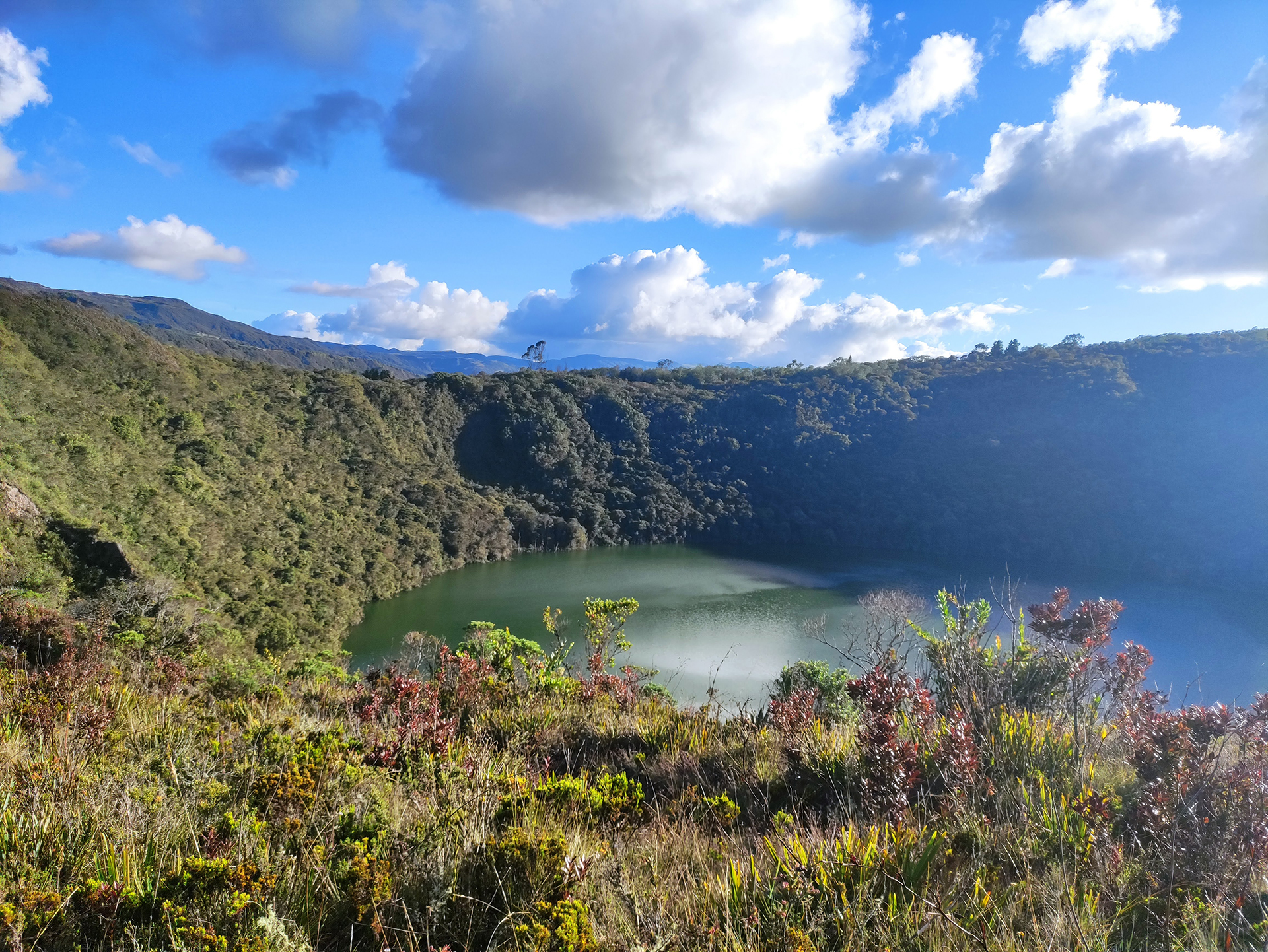 A crater lake surrounded by dense green vegetation under a partly cloudy blue sky, the landscape features rugged terrain and lush plant life—one of the must-see sights on tours in Bogotá.