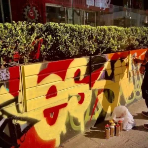 A person in a black shirt is spray-painting graffiti on a wall with yellow and red colors, next to a row of bushes. Several spray paint cans and a plastic bag are on the ground. Exploring urban art like this is one of the vibrant things to do on tours in Bogotá.