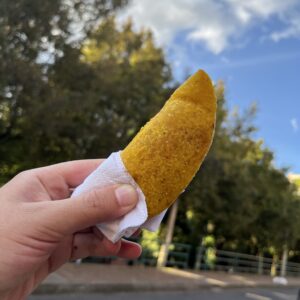 A hand holding a yellow empanada with a napkin against a backdrop of trees and a blue sky, perfectly capturing one of the delightful things to do in Bogotá.