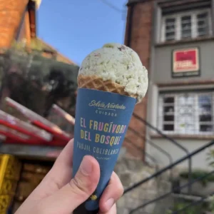 A hand holding an ice cream cone in front of a building. The cone has a blue wrapper with text in Spanish, including “El Frugívoro del Bosque.” It's one of those delightful things to do in Bogotá, making any tour of the city extra sweet.